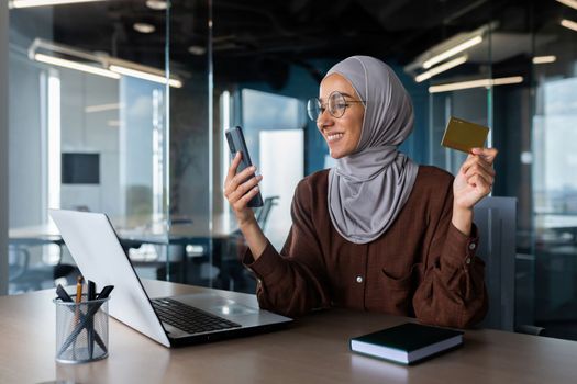 Successful businesswoman in hijab working inside office with laptop at workplace, muslim woman holding phone and bank credit card for online shopping and money transfer.