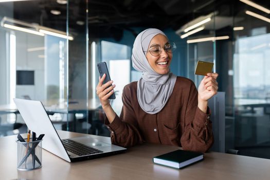 Successful businesswoman in hijab working inside office with laptop at workplace, muslim woman holding phone and bank credit card for online shopping and money transfer.