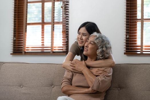 Loving adult daughter hugging older mother, standing behind couch at home, family enjoying tender moment together, young woman and mature mum or grandmother looking at each other, two generations..