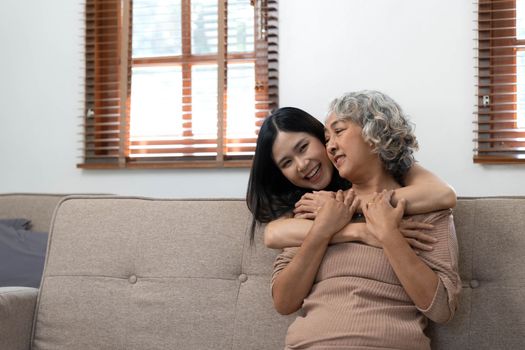 Loving adult daughter hugging older mother, standing behind couch at home, family enjoying tender moment together, young woman and mature mum or grandmother looking at each other, two generations..