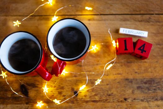 Two cups of coffee in red cups on a background of garlands. Postcard with date and month. Morning breakfast for Valentine's Day. Love concept