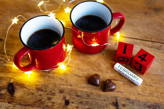 Two cups of coffee in red cups on a background of chocolate canfet. Postcard with date and month. Valentine's Day Morning Surprise