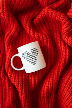 A white cup with a heart stands on a red knitted fabric. The concept of love and celebration of St. Valentine's day, February 14th