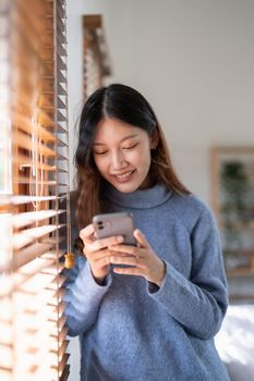 Cheerful young asian woman using mobile phone near window at home