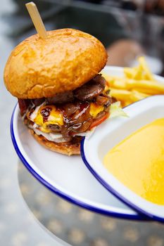 The concept of fast food and takeaway food. A juicy hamburger lies next to french fries on a metal plate along with cheddar cheese sauce in the background