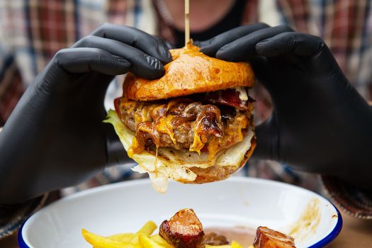 The concept of fast food and takeaway food. A man in black latex gloves holds a juicy hamburger in his hands, lies near french fries on a metal plate along with cheese sauce