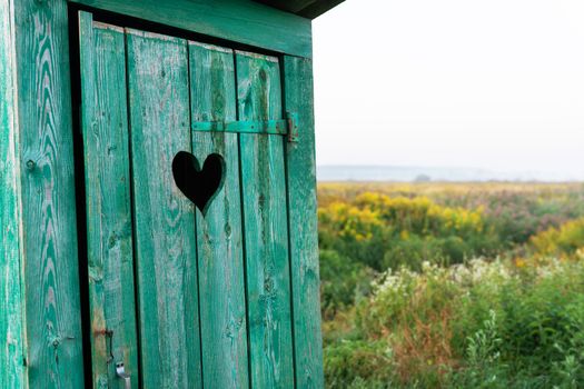 Heart shape on the old wooden toilet door, green toilet in the field. Outdoor recreation. Close-up