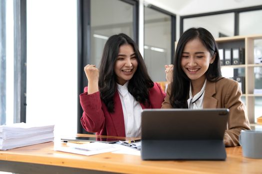 Two young Asian businesswomen show joyful expression of success at work smiling happily with a laptop computer in a modern office..