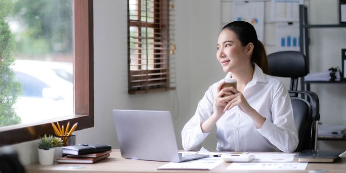 Beautiful young Asian businesswoman smiling holding a coffee mug and laptop working at the office...