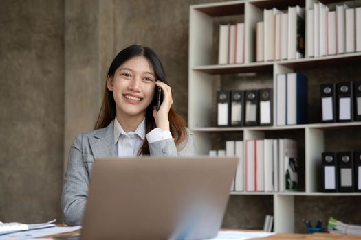 Smiling beautiful Asian businesswoman analyzing chart and graph showing changes on the market and holding smartphone at office...