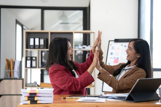 Two young Asian businesswomen show joyful expression of success at work smiling happily with a laptop computer in a modern office..