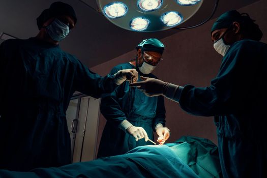 Surgical team performing surgery to patient in sterile operating room. In a surgery room lit by a lamp, a professional and confident surgical team provides medical care to an unconscious patient.