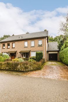 a brick house in the middle of a graveled driveway with trees and bushes around it on a cloudy day