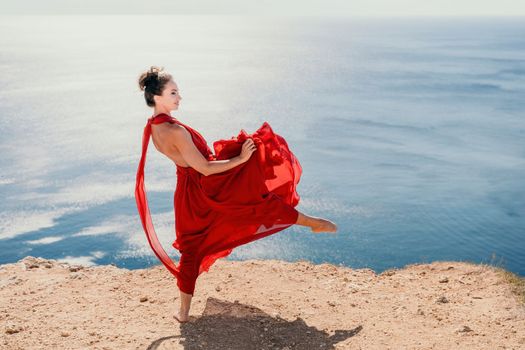 Side view a Young beautiful sensual woman in a red long dress posing on a rock high above the sea during sunrise. Girl on the nature on blue sky background. Fashion photo.