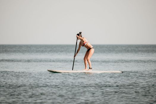 Sea woman sup. Silhouette of happy middle aged woman in rainbow bikini, surfing on SUP board, confident paddling through water surface. Idyllic sunset. Active lifestyle at sea or river