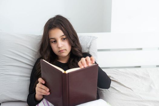 Caucasian teenage girl is lying on the bed and reading book. Reading fiction, hobbies, leisure activities and recreation. Concept of home education.