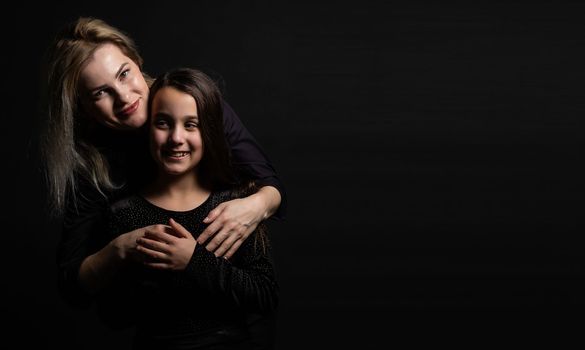 Young mother and her little daughter are looking at camera and smiling, sitting against black wall.