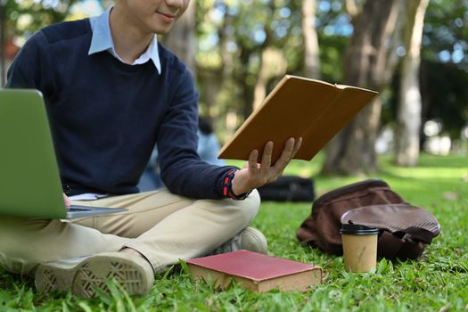 Cropped shot of male college student using laptop on green meadow in campus. Education, youth and technology concept.