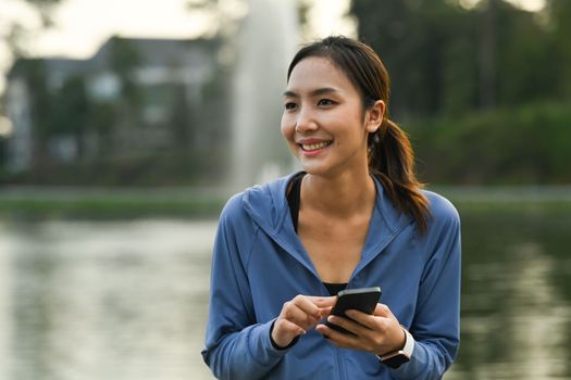 Charming asian female runner in sport clothes using smart phone during morning workout in the park.