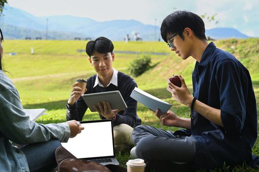 Group of young college students reading book on green lawn at campus. Youth lifestyle, university and friendship concept.