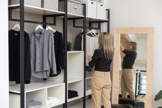 Woman choosing outfit from large wardrobe closet with stylish clothes and home stuff