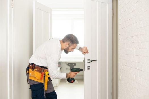 Installation of a lock on the front wooden entrance door. Portrait of young locksmith workman in blue uniform installing door knob. Professional repair service. Maintenance Concept.