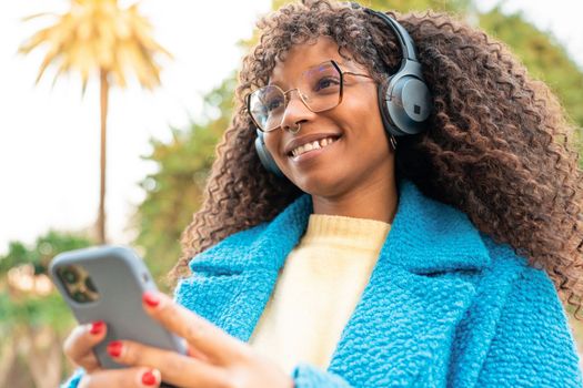 Happy African American woman using headphones and smart phone enjoying music, podcast our audio books outdoors in the park. High quality photo