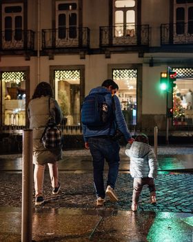 father holds hand of little daughter and family crossing road at night with the green traffic light.