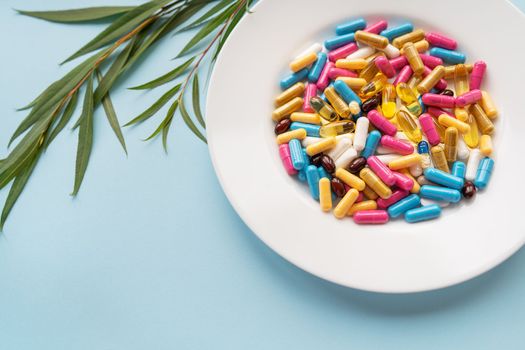 White plate with pills of nutritional supplements in different bright colors. Blue background, place for an inscription. Green branch on the background