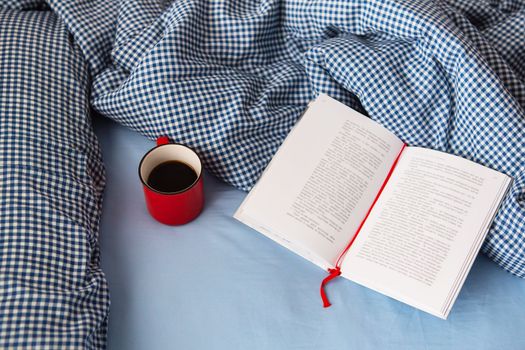 Ukraine, Rivne - August 11, 2021: Top view of a cozy blanket, an open book and a hot red cup of coffee on the bed on a cold winter day. Relaxation and hygge concept. Top view with copy space