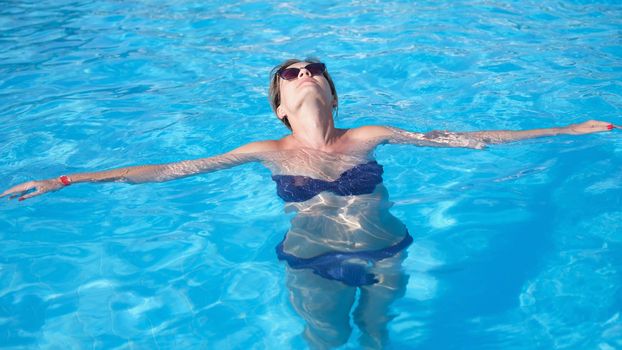 Glamorous woman in stylish sunglasses enjoys swimming in turquoise water in pool. Relaxing in pool of luxury resort