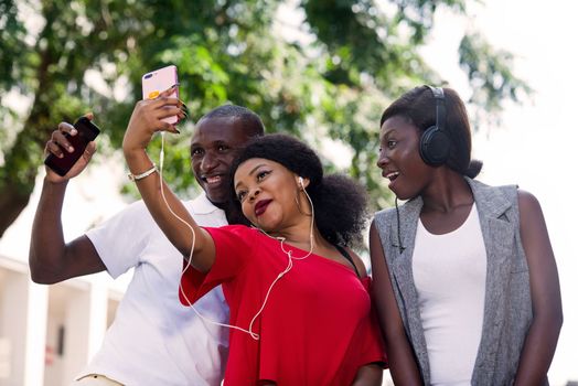 group of three happy young people with mobile phone takes picture and listens to music with headphone outside