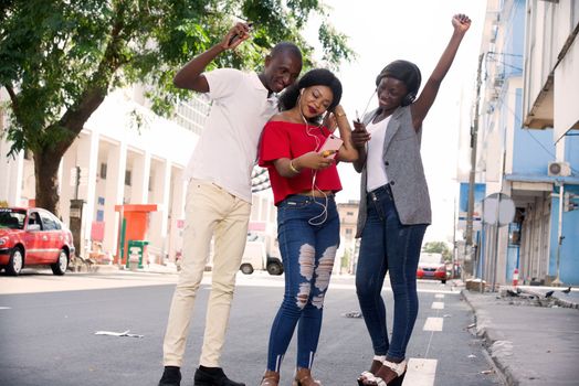 group of three happy young people with mobile phone takes picture and listens to music with headphone outside