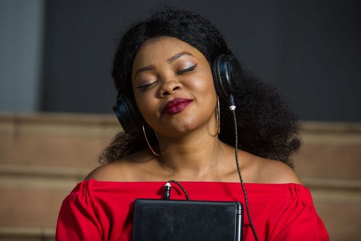 young girl sitting in red camisole with laptop listening to music using headphones with eyes closed while smiling.