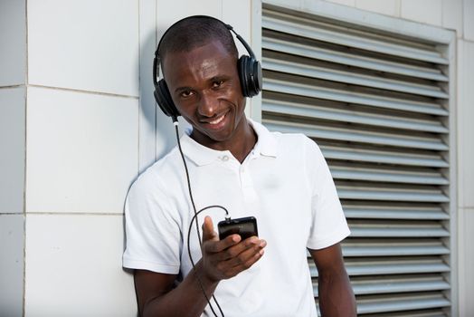 people, music, technology, leisure and lifestyle - Happy african young man with headphones and smart phone on the street