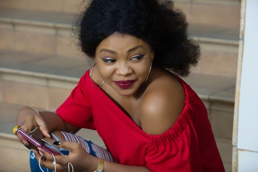 Funny african woman sitting on stairs listening to music with headphones and a smart phone outdoors.