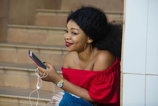 Funny african woman sitting on stairs listening to music with headphones and a smart phone outdoors.