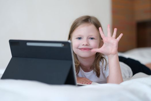 Portrait of a smiling child girl with tablet on bed holds hand in greeting. Gadgets for development and games in children