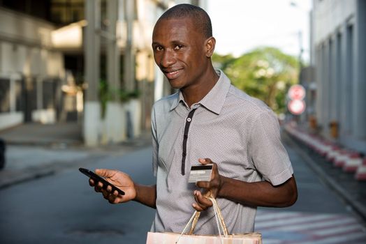 young man holding shopping bags and credit cards while handling a mobile phone outdoors. Shopping and fashion concept.