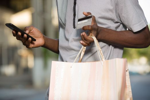 young man holding shopping bags and credit cards while handling a mobile phone outdoors. Shopping and fashion concept.