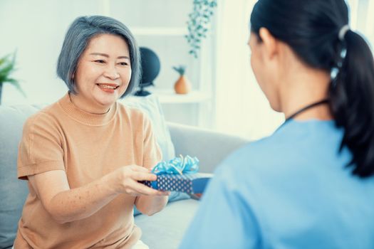 A young caregiver hand over to her senior patient a blue gift box with blue ribbons at a contented living room.