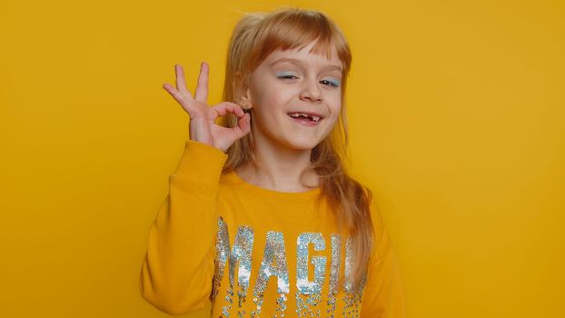 Ok. Young toddler school girl in sweater looking approvingly at camera showing okey gesture, like sign positive, approve something good. Teenager female child kid isolated on studio yellow background