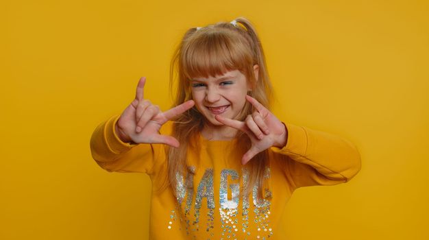 Overjoyed happy young child girl kid showing rock n roll gesture by hands, cool sign, shouting yeah with crazy expression, dancing, rejoicing in success. Little toddler children on yellow background