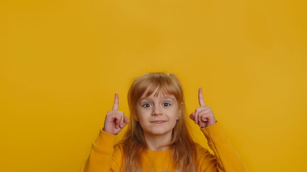 Happy toddler girl showing thumbs up pointing empty place, advertising area for commercial text, copy space for goods promotion banner. Young little child kid. Studio shot indoors on yellow background