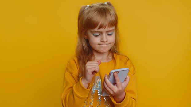 Young toddler school girl in sweater use smartphone typing browsing, loses becoming surprised sudden lottery results, bad fortune, loss. Education, study. Teen female child kid on yellow background