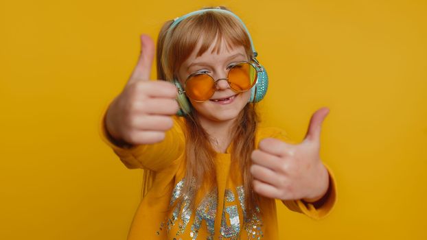 Happy preteen child girl kid listening music via headphones and dancing disco fooling around having fun expressive gesticulating hands. Little toddler children isolated on studio yellow background