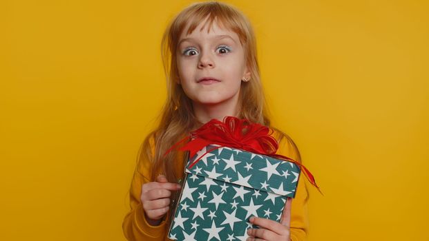 Lovely smiling young preteen child girl kid presenting birthday gift box stretches out hands, offer wrapped present career bonus, celebrating party. Little toddler children on studio yellow background