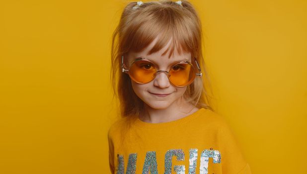 Smiling playful happy young preteen child girl kid walking slowly and looking at camera, flirting. Little charismatic lovely toddler children in orange sunglasses isolated on yellow wall background