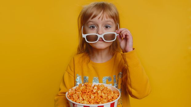 Excited young toddler school girl in 3D glasses eating popcorn, watching interesting tv serial, sport game, film, online social media movie content. Teen female child kid on studio yellow background