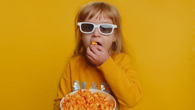 Excited young toddler school girl in 3D glasses eating popcorn, watching interesting tv serial, sport game, film, online social media movie content. Teen female child kid on studio yellow background